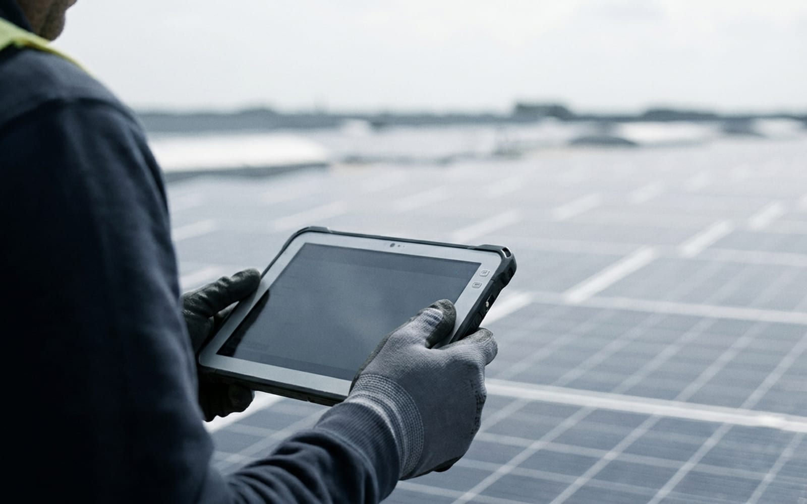 Le mani di un tecnico che tengono un tablet robusto sul tetto di un capannone coperto di pannelli fotovoltaici.
