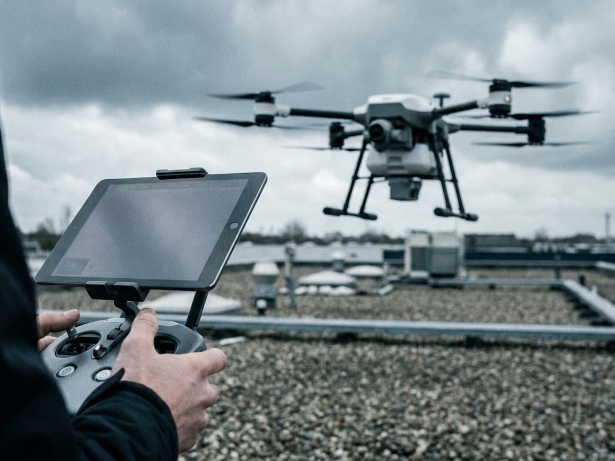 Le mani di un operatore sul radiocomando con tablet agganciato, mentre un drone vola davanti a lui sopra il tetto di un capannone.